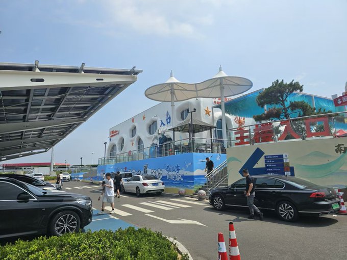 An outdoor daytime scene at a Chinese tourist attraction with cartoon-star decorations on a white building, a pedestrian crosswalk, parked cars, and pedestrians walking near tents and traffic cones.