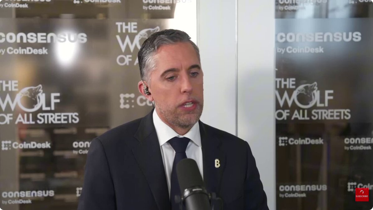 The image features a man dressed in a suit, standing at a microphone and speaking into it. He appears to be addressing an audience or participating in a press conference. The background includes a large window with a white frame, which has "The Wolf of All Streets" written on it.