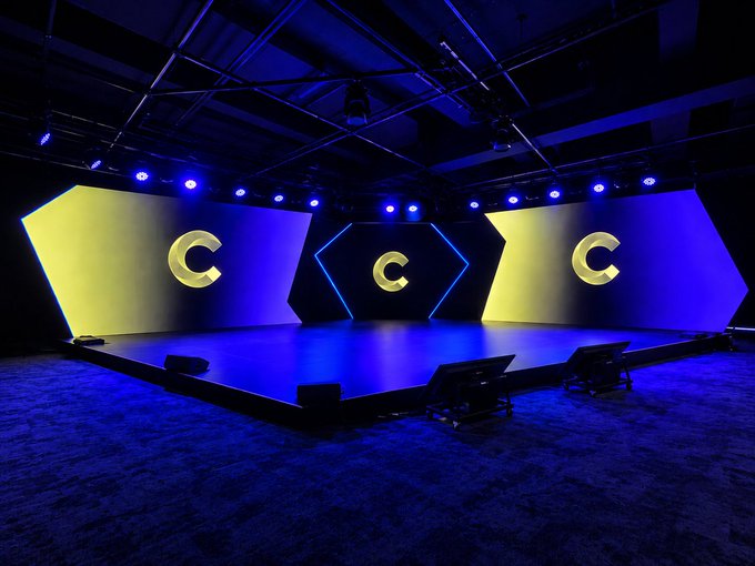 A modern stage setup with blue lighting, featuring three large screens displaying a yellow 'C' logo against blue and yellow backdrops.