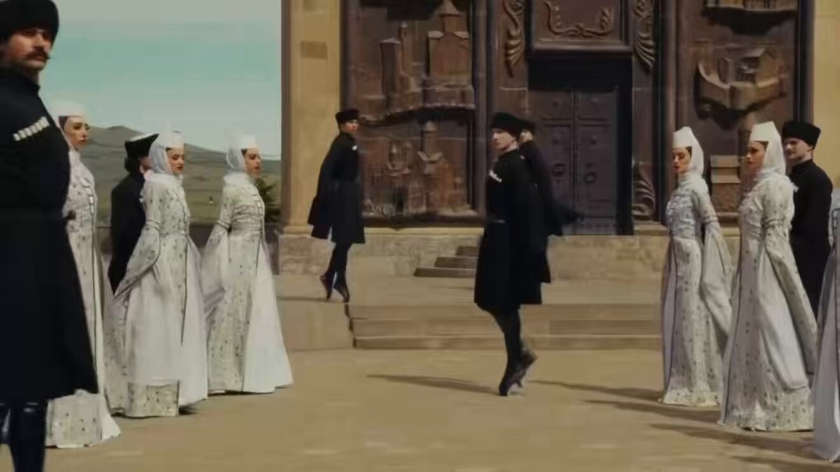 The image features a group of people dressed in white robes, standing outside an old building with a large wooden door. The scene appears to be set in a museum or gallery setting, as suggested by the presence of a painting hanging above them. One person is seen walking towards the group, possibly joining their ranks as a member of the fine art community.