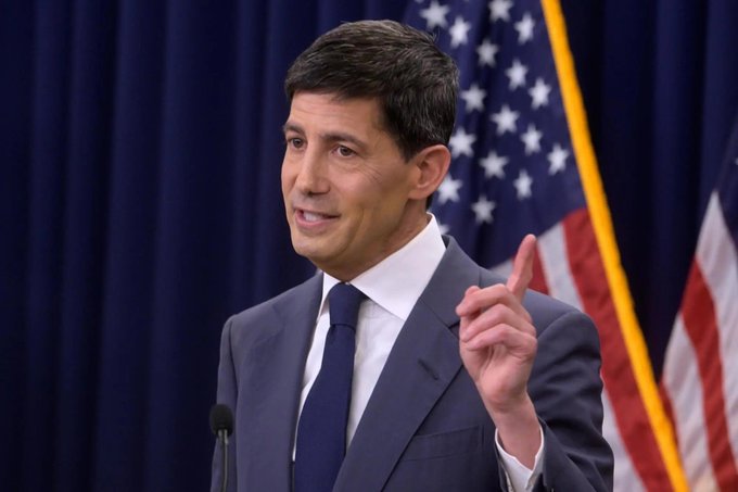 The image shows Fed Chair Kevin Warsh speaking at a podium with an American flag in the background, gesturing with his hand as he addresses an audience. This relates to the post as it highlights his role in releasing his first FOMC Minutes at 11:30 PM IST.
