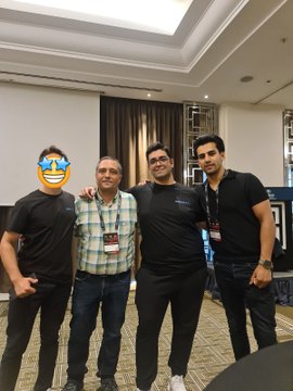 Four men pose together at what appears to be an indoor event, with one person's face covered by a laughing emoji sticker; they wear lanyards suggesting a conference setting.