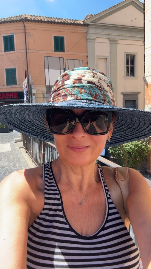 A selfie of a woman wearing a large patterned sun hat, dark sunglasses, and a striped tank top, with orange and beige European-style buildings visible behind her on a sunny day.