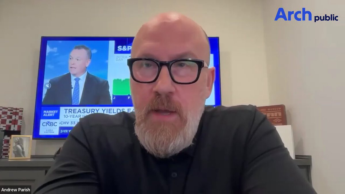 The image shows an older man wearing glasses, sitting at a desk with a TV screen behind him displaying two news anchors talking about Treasury yields. The man appears to be engaged in a conversation or presentation related to this topic.