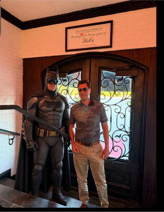 A man in a patterned polo shirt and khaki pants posing next to a Batman statue at Walt's, with a sign reading 'THE VINTAGE BURGER CO. thanks you for being a part of Walt's' above the doorway.