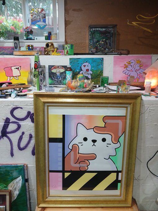 An artist's studio featuring a framed painting of a cat with a pointed finger, displayed on a ledge surrounded by other colorful artworks, paint supplies, and decorative objects.