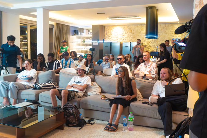 A group of casually dressed people sit on sofas in a modern living room, watching a presentation while being filmed by a professional camera operator on the right.