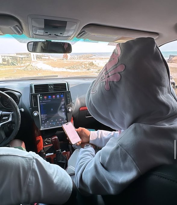 An interior view of a car showing a person wearing a gray hoodie holding a smartphone, with the driver's dashboard, navigation screen, and a glimpse of the sea visible through the windshield.