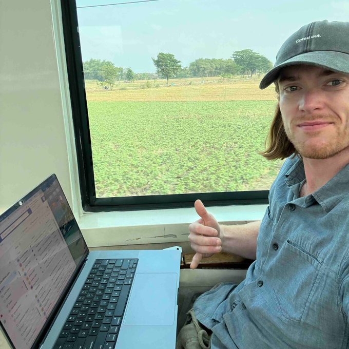 A man with shoulder-length hair and a gray "Clothed dust" cap gives a thumbs-up while sitting by an open laptop, with green fields visible through the window behind him.