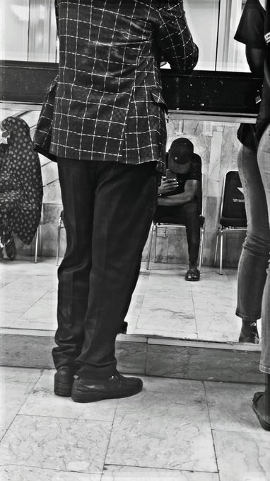A black-and-white photo showing a person in a checkered blazer and dark pants standing in a lobby, with another person seated on a chair looking at their phone in the background.