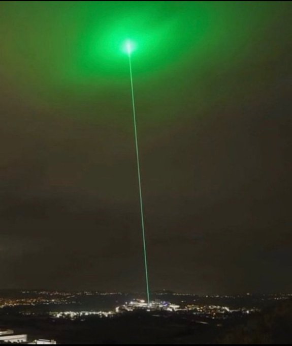 A nighttime photograph showing a bright green laser beam shooting up into the sky from a city below, illuminating clouds with a vivid green glow.