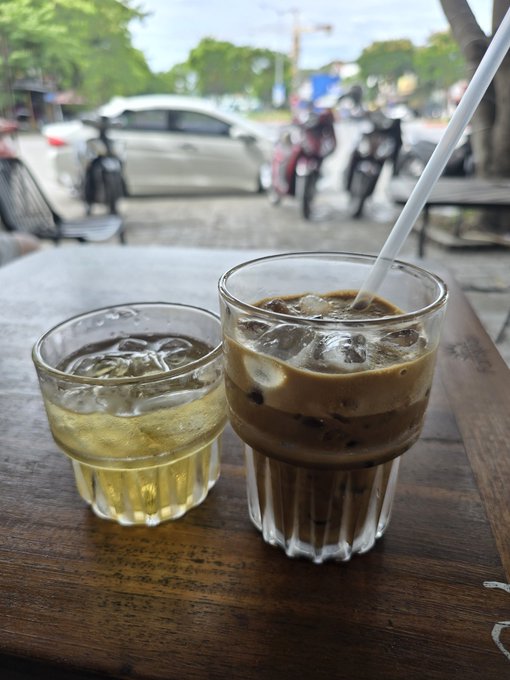 Two iced beverages in glass cups on a wooden table at a sidewalk cafe, with parked motorcycles visible in the background.