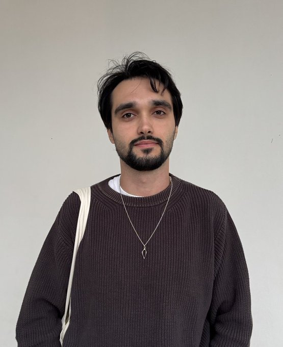 A portrait of a man with dark hair and a beard, wearing a brown knit sweater over a white shirt and a pendant necklace.