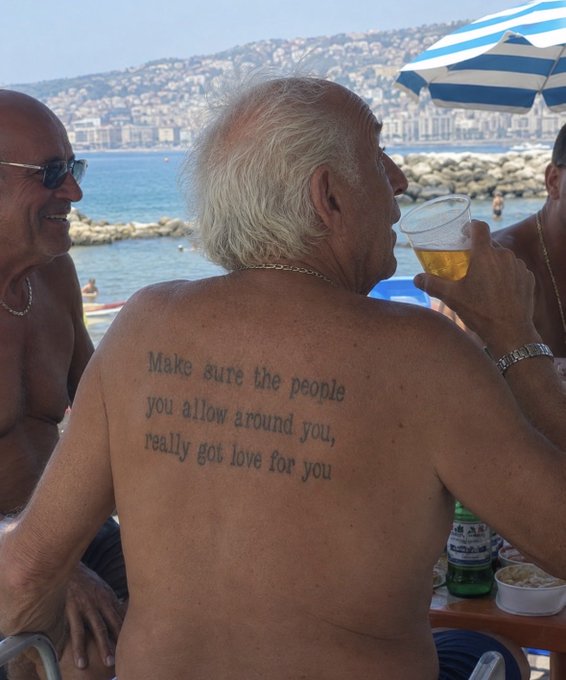 An older man with white hair drinks beer at a beach, showing a back tattoo that reads, 'Make sure the people you allow around you, really got love for you'.
