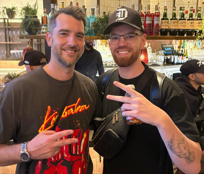 Two smiling men pose together at a bar, with one wearing a "Detroit" cap and a black shirt while making a peace sign, and the other in a "Bá Bá" graphic tee also flashing a peace sign. The bar's well-stocked shelves featuring Campari and other liquor bottles serve as a vibrant backdrop, capturing a fun social moment that aligns with the @doginaldogs community's celebratory vibe referenced in the post.