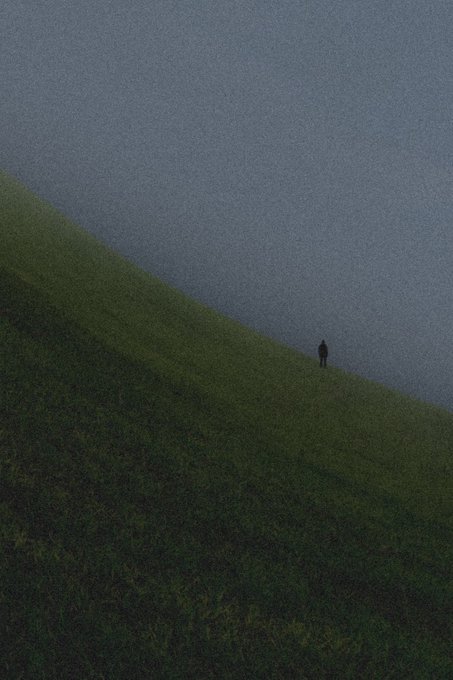 A minimalist photograph shows a small lone figure walking along a diagonal ridge where green grass meets a gray textured surface, creating a stark atmospheric composition.