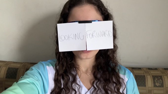 A woman with long curly hair holds a handwritten sign reading 'LOOKING FORWARD' over her eyes while sitting on a patterned couch.