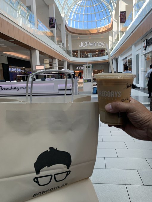 A hand holds an iced coffee next to a shopping bag with a glasses-wearing face logo and '#GREGCULAR', taken inside a mall with a JCPenney store visible in the background.