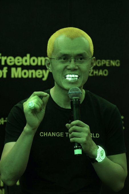 A man with yellow hair and face paint featuring a green hue smiles while speaking into a microphone, wearing a black t-shirt reading 'CHANGE THE WORLD' at a Freedom of Money event.