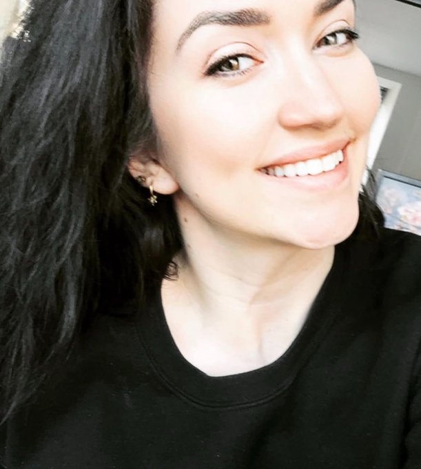 A close-up selfie of a smiling woman with long dark hair wearing a black shirt and small gold earrings.
