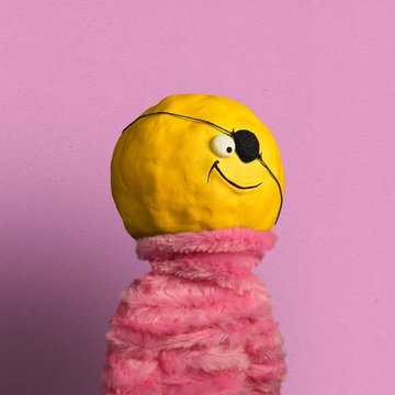 A craft figure made of clay with a yellow round head featuring a simple smiley face and a body covered in pink fluffy material, set against a pink background.