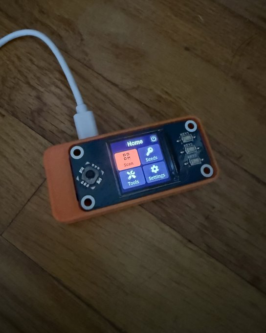 A small electronic device with a USB-C cable resting on a wooden surface, featuring a color display showing a 'Home' menu with Scan, Seeds, Tools, and Settings options, housed in a 3D-printed orange case.