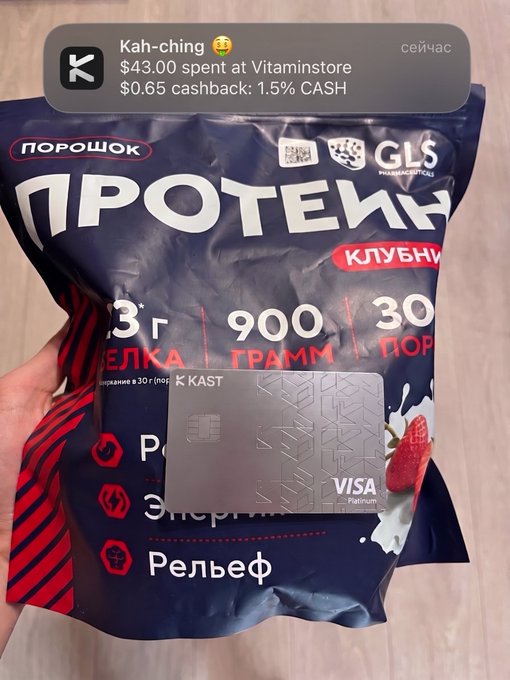 A hand holds a 900g bag of strawberry-flavored protein powder (Протеин, клубника) from Vitaminstore alongside a silver KAST Visa Platinum card. An overlay notification shows a $43.00 purchase earning $0.65 (1.5%) cashback instantly, illustrating the post's point about using crypto for everyday spending.
