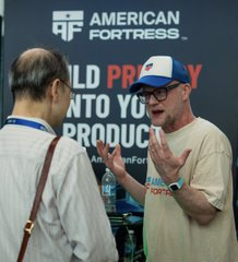 A man in a white and blue trucker hat, glasses, and a beige t-shirt gestures while speaking to another man at an 'AMERICAN FORTRESS' booth.