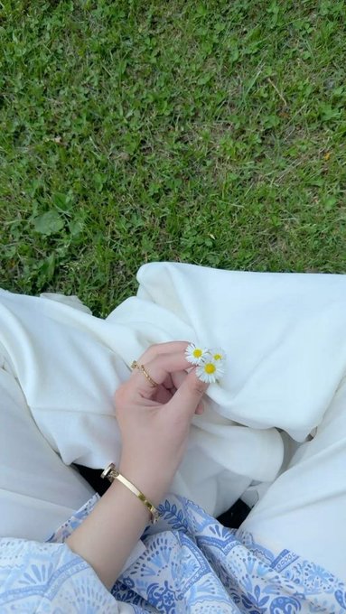 A person's hand wearing gold rings and a bracelet, holding two small daisies while sitting on grass with a white garment draped over their lap.