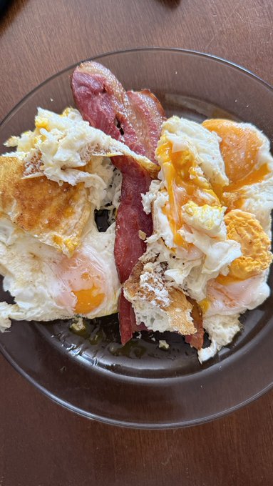 A brown bowl filled with a hearty breakfast of fried eggs with runny yolks, crispy bacon, and toasted bread slices on a wooden table.