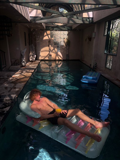 A shirtless man with blonde hair lounges on a colorful inflatable raft in a sunlit indoor pool, holding a martini glass while relaxed against a built-in headrest, with another inflatable float visible in the background and sunlight streaming through skylights onto the pool's tiled floor.