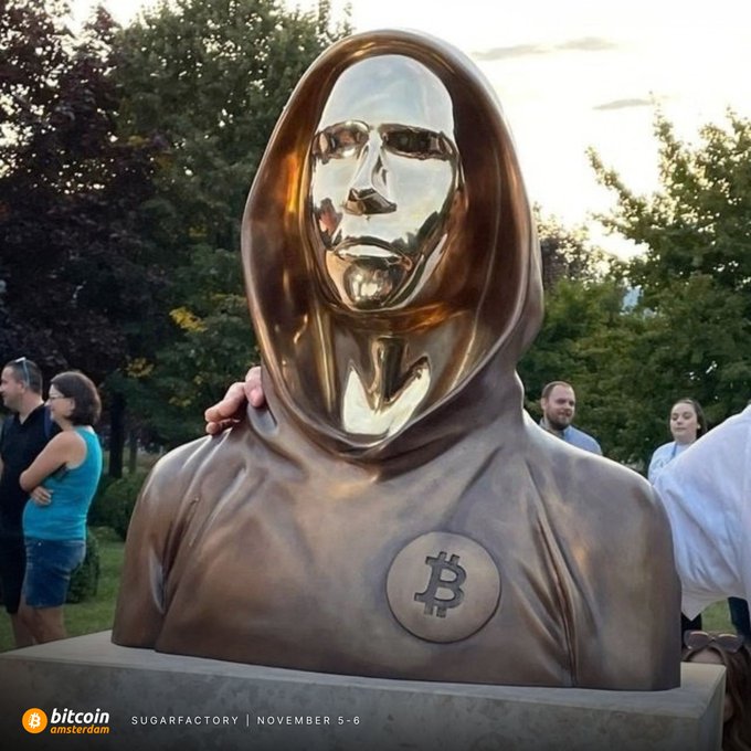 A reflective bronze bust of Satoshi Nakamoto, hooded and bearing the Bitcoin logo on its chest, is unveiled at SugarFactory as onlookers gather around. The image ties to the post's message that the mirrored face symbolizes "we are all Satoshi," highlighting the communal ethos behind Bitcoin Amsterdam on November 5–6.