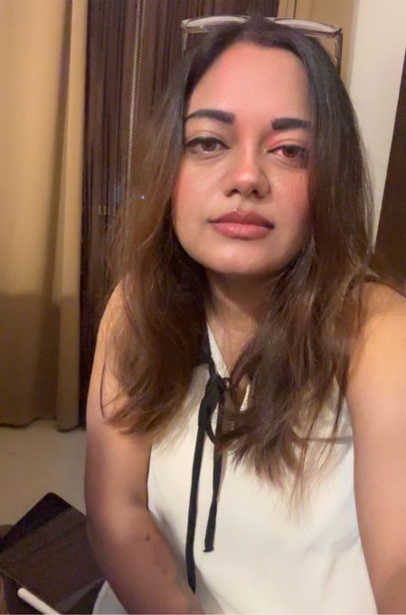 A selfie of a woman with long brown hair wearing a white sleeveless top, with glasses resting on her head.