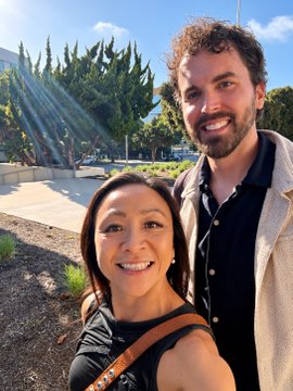 A selfie of a smiling Asian woman with brown hair and a bearded man with curly hair standing outdoors in front of trees and a building.