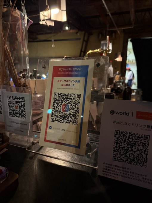 A close-up of payment QR code signs displayed at a bar or restaurant counter in Okayama, including a HeartPort Wallet sign promoting "JPYC" (a stablecoin pegged 1:1 to the Japanese yen) and a World ID code, illustrating the growing adoption of crypto payments alongside traditional options like cash.