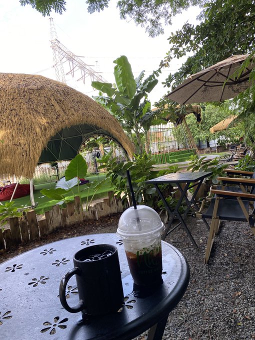 A black ceramic coffee mug and a plastic cup of iced coffee with a straw sit on a decorative metal table at an outdoor café, with a thatched-roof hut and tropical plants in the background, perfectly illustrating the relaxed, contented vibe of someone who bought Bitcoin without worrying about the cost basis.