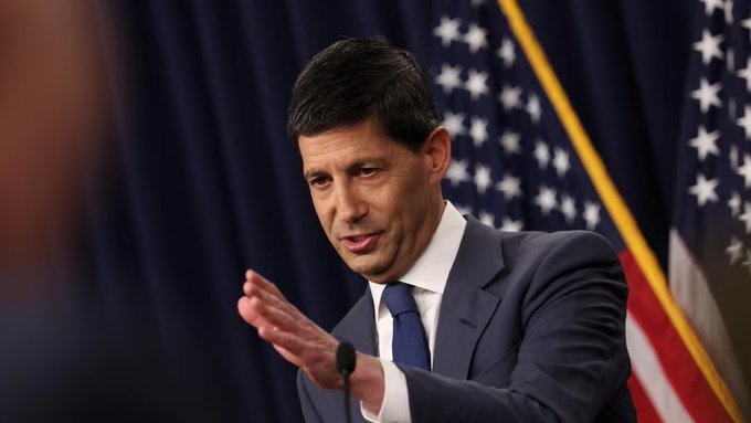 A man in a suit gesturing while speaking in front of a US flag, appearing to be at a formal event or press conference.