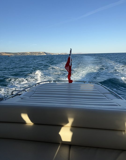 The rear deck of a boat cuts through open blue water, flying a British Red Ensign flag, with a distant coastline visible ahead. The image pairs with the post's message of enjoying a leisurely weekend on the south coast while Bitcoin's price remains flat.
