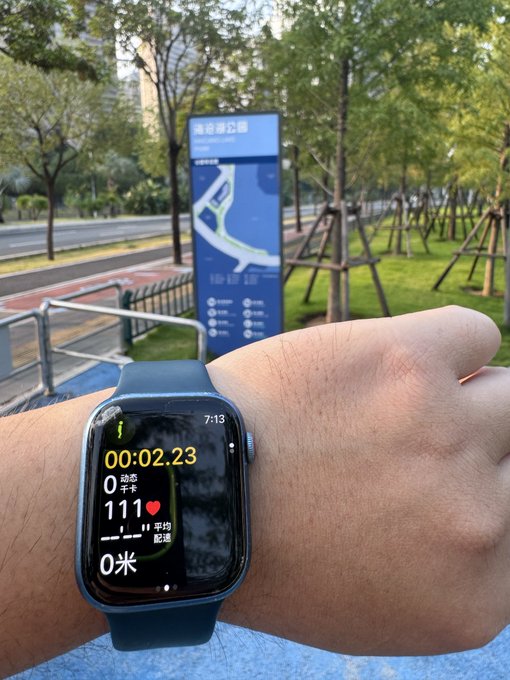 A wrist wearing an Apple Watch displays a workout in progress with a timer at 00:02.23, a heart rate of 111 BPM, and 0 meters, set against the backdrop of a park and an exercise station sign.