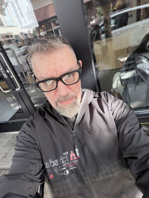 A middle-aged man with gray hair, a beard, and black glasses takes a selfie while standing outside, wearing a dark jacket with 'lab nctu' text.