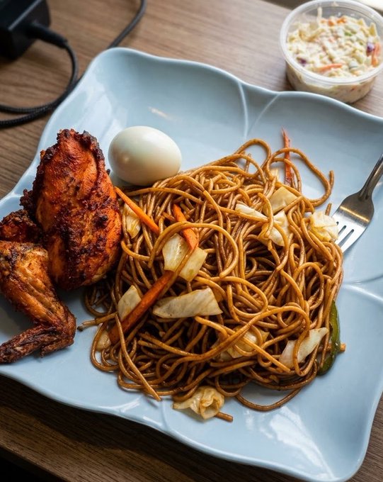 A plate of stir-fried noodles served with fried chicken, a boiled egg, and vegetables, accompanied by a side of coleslaw on a wooden table.