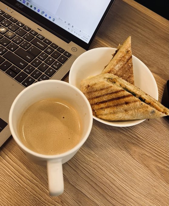 A photo of a laptop next to a white mug of coffee and a paper plate with grilled sandwich triangles on a wooden desk.