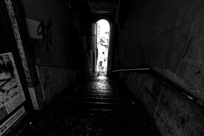 A black and white photograph looking down a dark, graffiti-covered staircase toward a bright, arched exit leading to a sunlit European street.