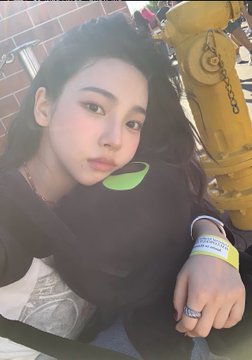 A young woman with long dark hair wearing a black top and a neon green wristband takes a selfie in front of a brick wall and yellow construction equipment.