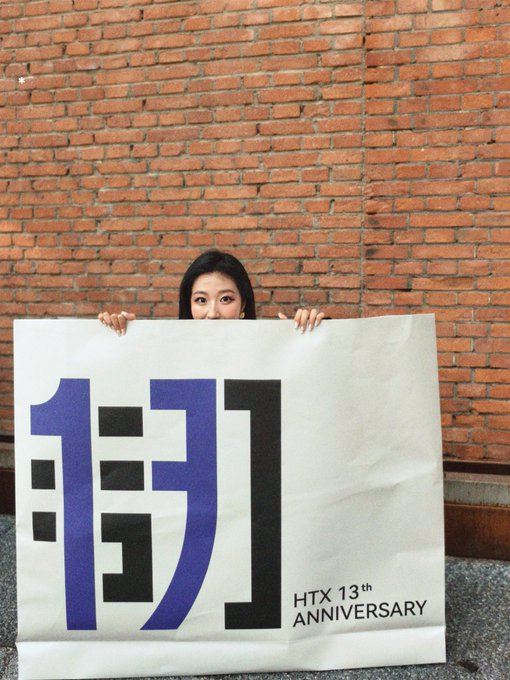 A woman peeks over a large white bag featuring bold blue Chinese characters "倒闭" (bankruptcy) alongside "HTX 13th ANNIVERSARY" text, standing against a brick wall—visually contrasting the celebratory anniversary merchandise with an ironic message about HTX's struggles.