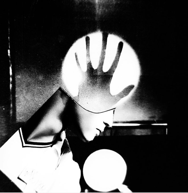 A dramatic black and white surreal photograph showing a figure in a suit and hat with their hand raised against a glowing circular light, creating a strong contrast.