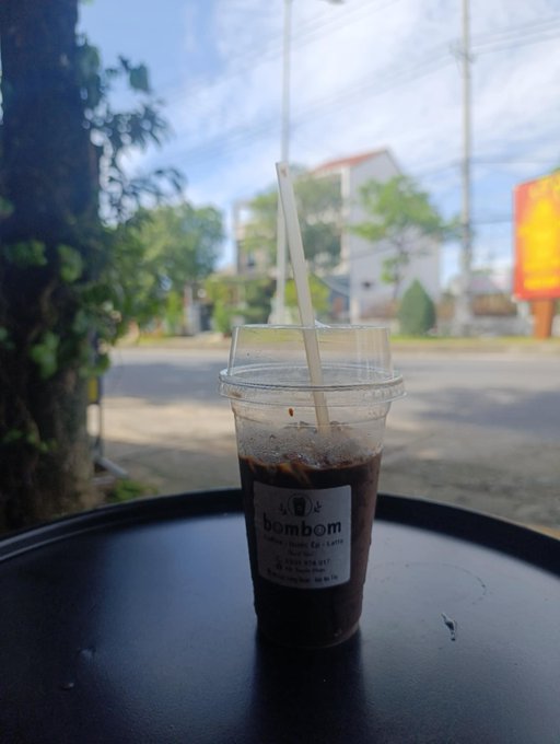 A plastic cup of iced coffee from 'bombom Coffee' shop sitting on a black tray/table with a straw, photographed from inside a shop looking out onto a street scene.