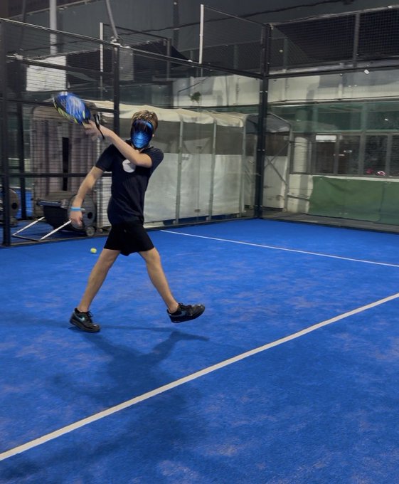 A young player swings a paddle tennis racket mid-swing on an indoor blue court in Singapore, with a bright yellow ball bouncing nearby beneath the glass-enclosed arena lights.