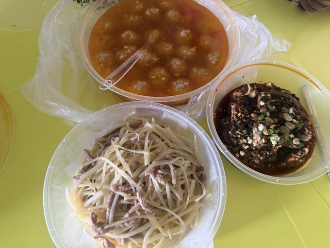 Three plastic containers of food on a yellow table: one with meatballs in tomato sauce, one with shredded vegetables and meat, and one with a dark dish topped with green onions.