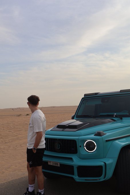A young man in a white tee and black shorts stands beside a striking turquoise Brabus G-Wagon (license plate 31055) parked in the desert, embodying the "GM (get money)" mentality with a high-end luxury vehicle as a status symbol of financial success.